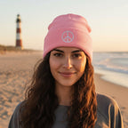 A light pink knit beanie hat with a folded cuff, featuring a white embroidered peace sign on the front.