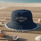 A navy blue bucket hat with a frayed white edge and 'OLD SHELLER' over a junonia seashell embroidered on the front, resting on a wooden boardwalk with seashells and a beach in the background.