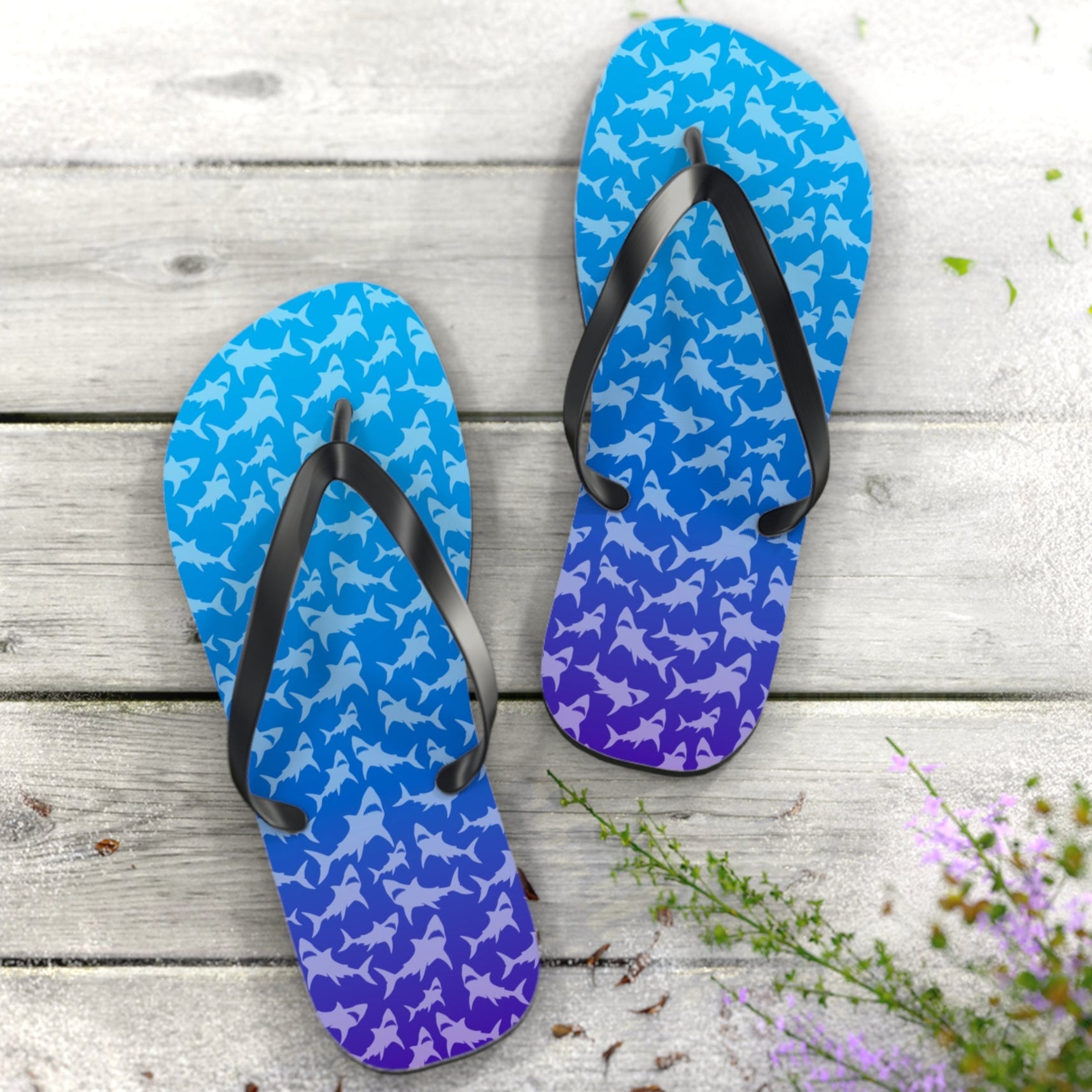Blue and purple ombré sharks pattern flip-flops on a wooden surface with flowers.