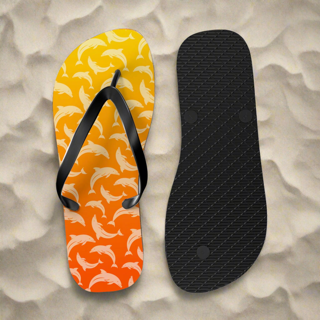 Flip-flop with dolphin pattern on a yellow and orange background, showing sole texture on a white beach background.