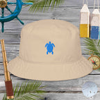 Beige bucket hat with blue turtle design on a wooden surface with nautical items.