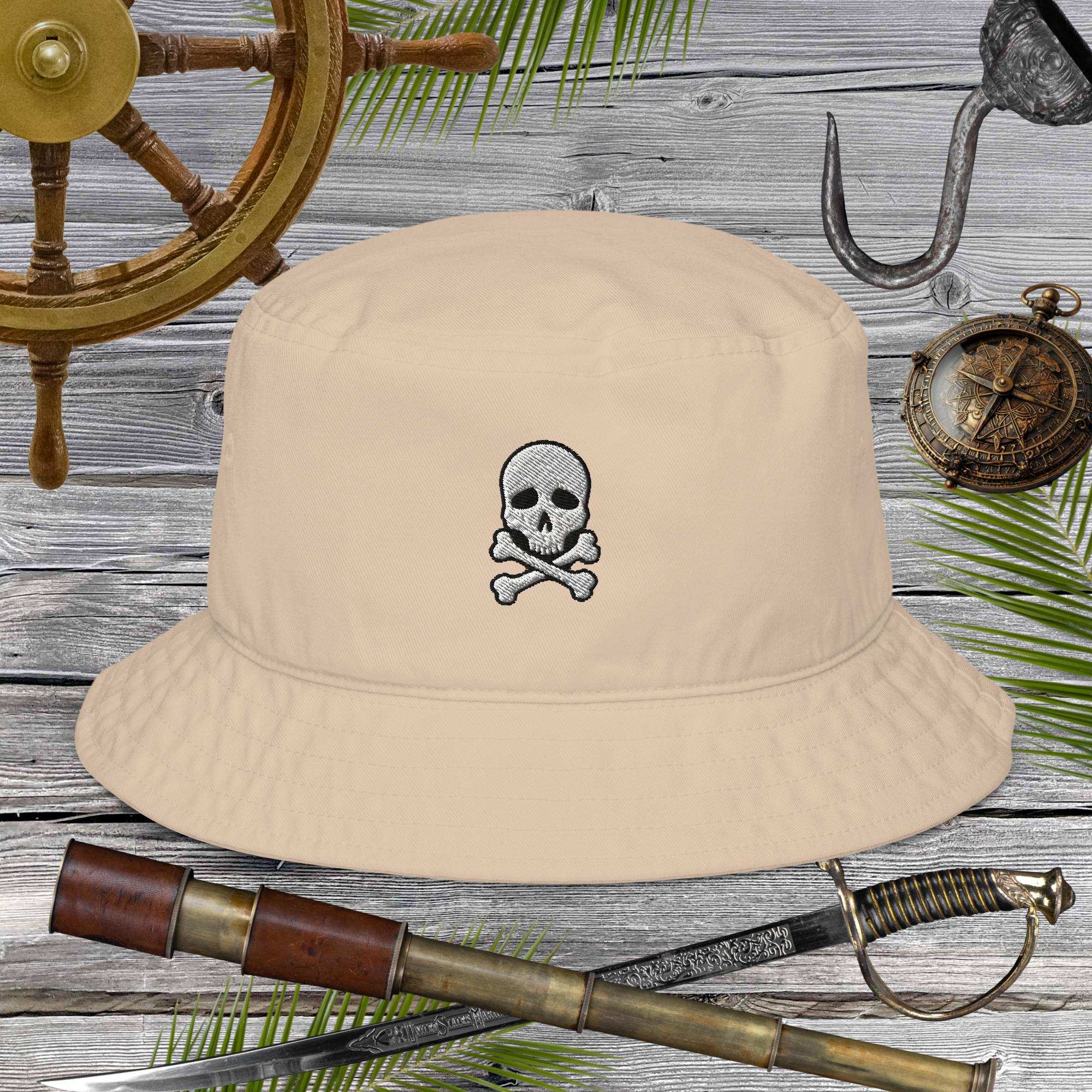 Beige bucket hat with skull and crossbones design on a wooden background with pirate-themed items.
