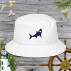 White bucket hat with shark embroidery on a wooden background with ship wheel and lighthouse.