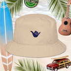 Beige bucket hat with embroidered blue shaka sign, surrounded by a surfboard, ukulele, and vintage car on a tropical background