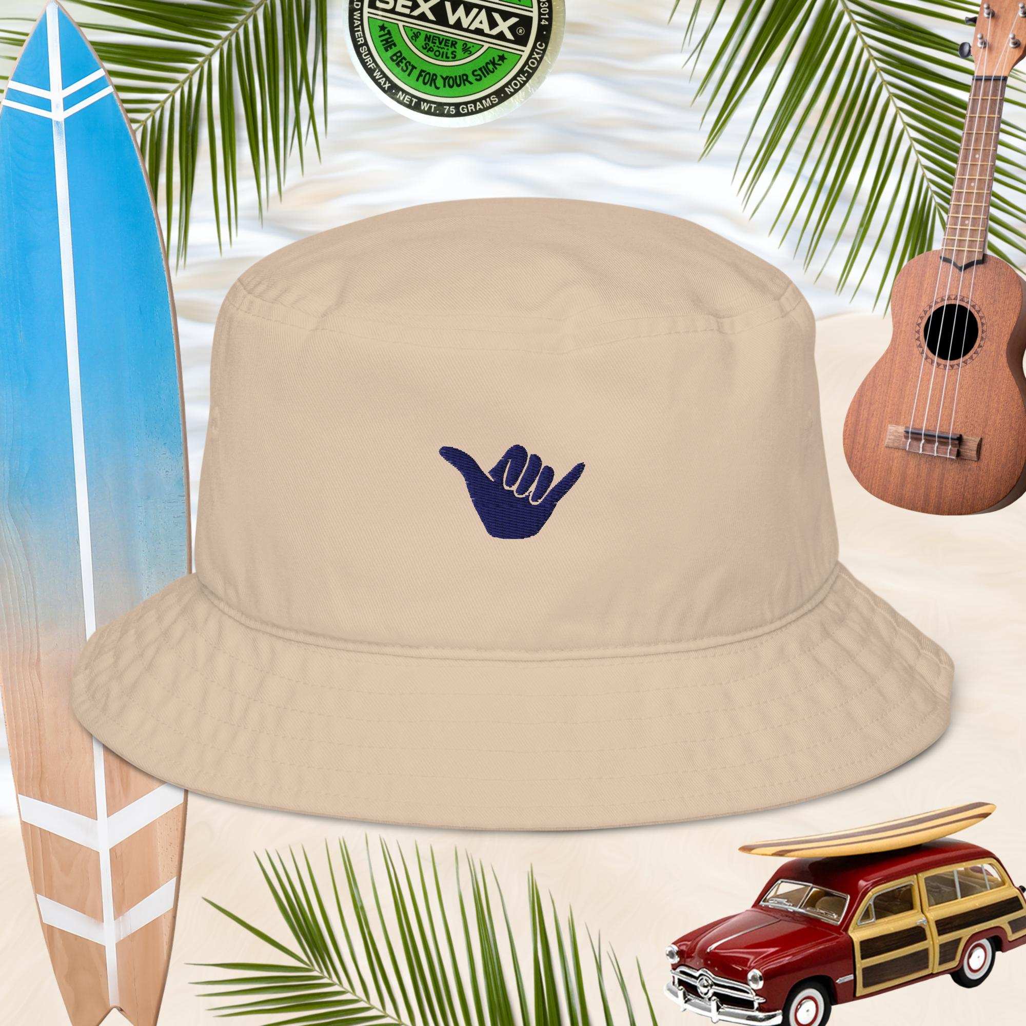 Beige bucket hat with embroidered blue shaka sign, surrounded by a surfboard, ukulele, and vintage car on a tropical background