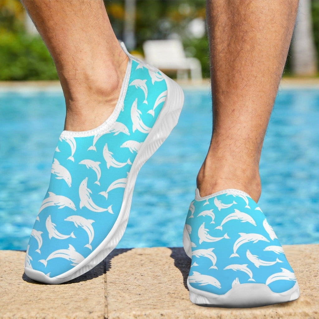 A person wearing slip-on water shoes with a light blue and white dolphin pattern, standing on a pool deck with a swimming pool and outdoor background visible.