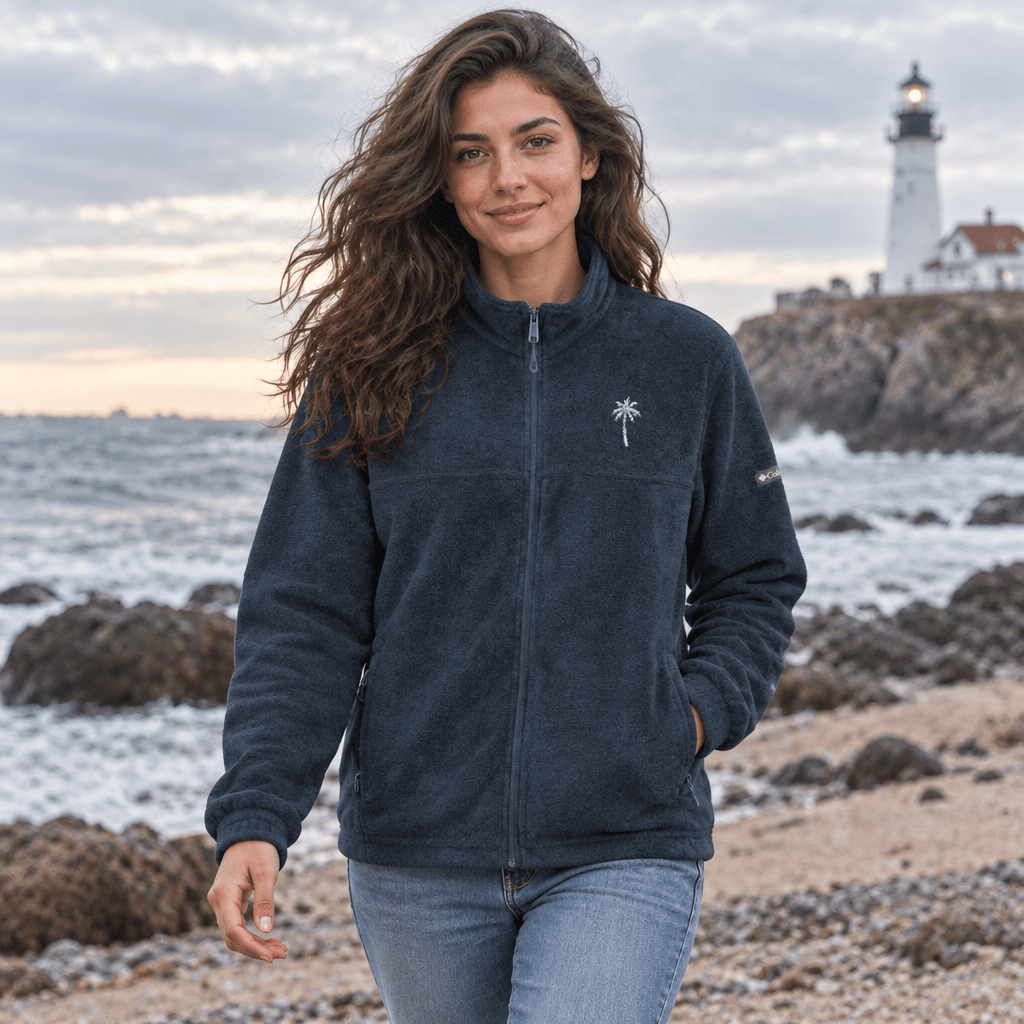 A woman stands on a rocky beach wearing a dark charcoal fleece zip-up jacket with a small white palm tree logo on the left chest, paired with light blue jeans. A lighthouse is visible in the background under a cloudy sky.