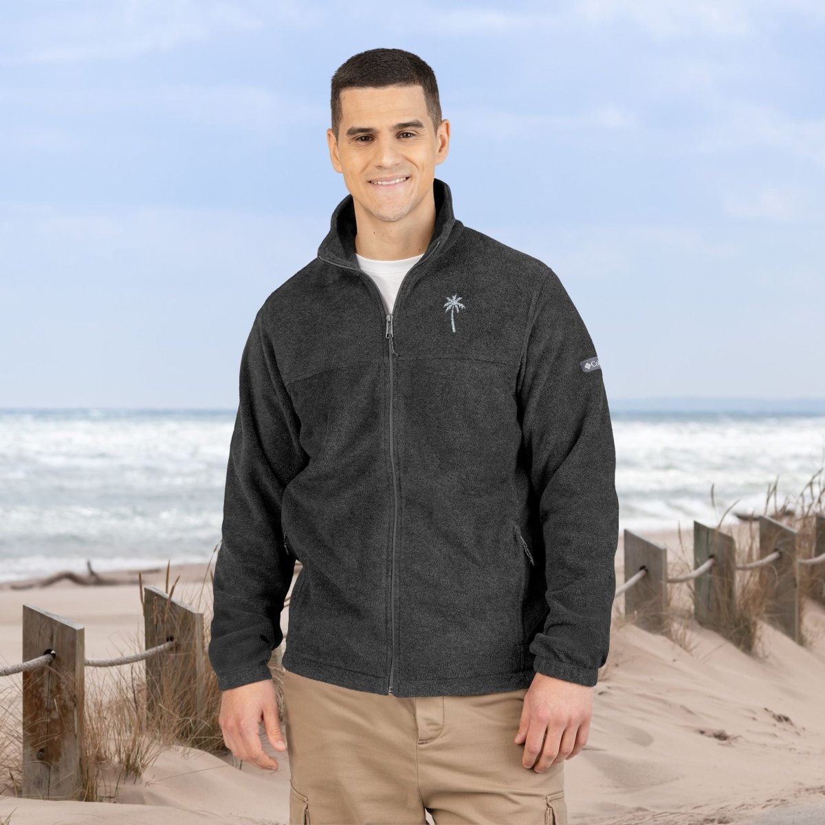 A man standing on a sandy beach wearing a dark charcoal gray zip-up fleece jacket with a white circular logo on the left chest, paired with khaki pants.