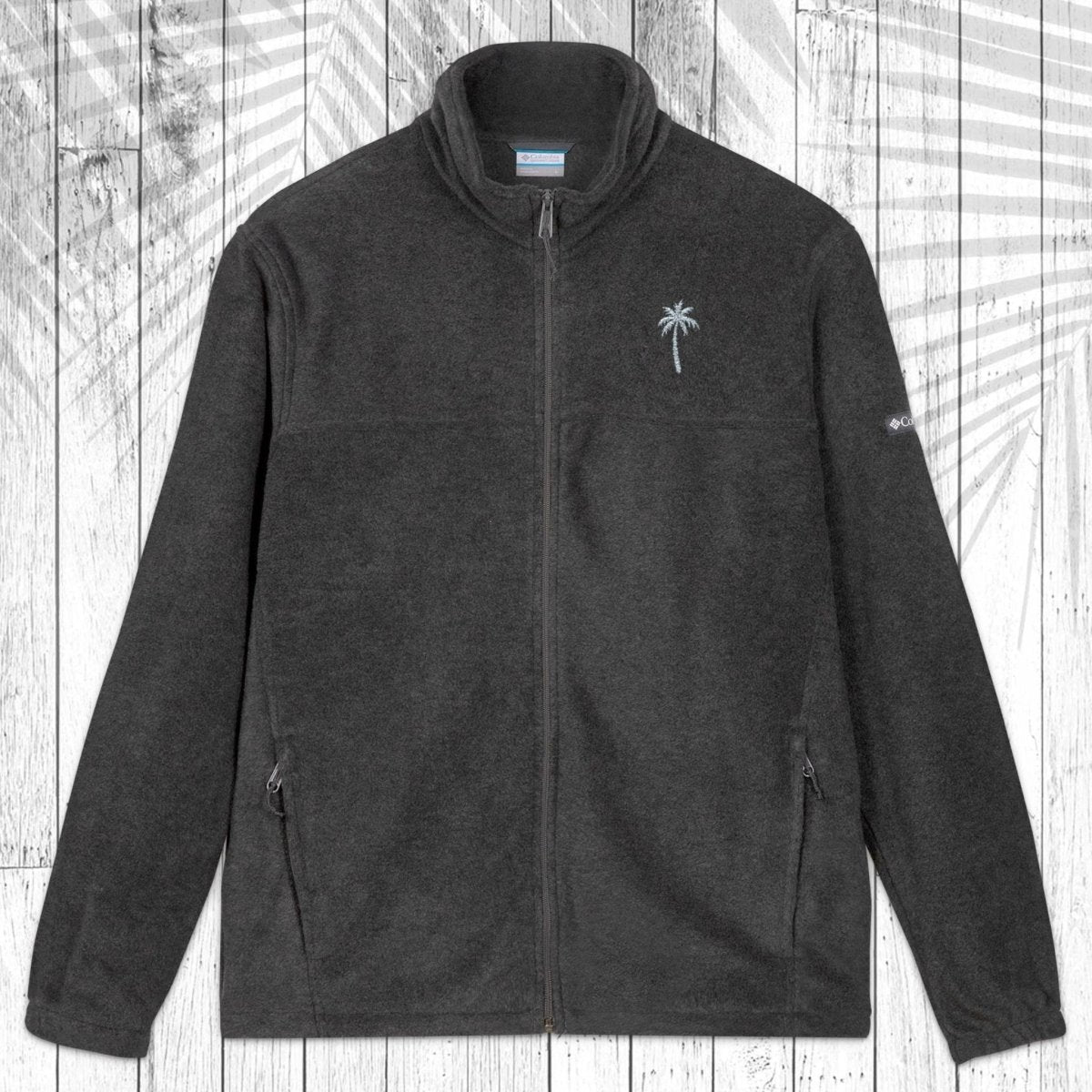 A dark gray fleece zip-up jacket with a stand collar, shown flat against a white wood plank background with a palm leaf pattern overlay. The jacket features a small embroidered  palm tree on the left chest and a small Columbia logo on the right sleeve.