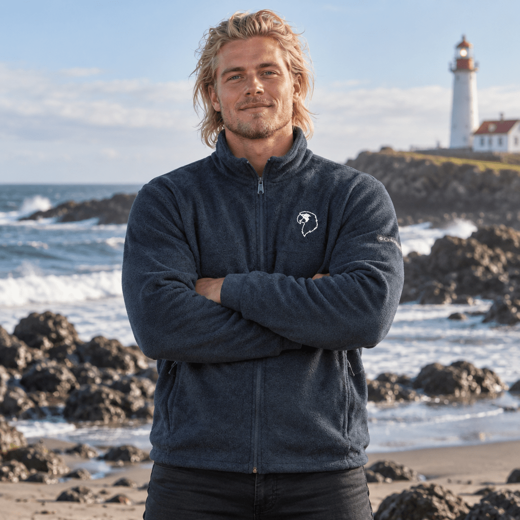 A man with blonde hair stands on a rocky beach with his arms crossed, wearing a dark gray fleece zip-up jacket with a small white logo on the left chest. In the background, there is an ocean scene with waves, rocks, and a lighthouse under a cloudy sky.
