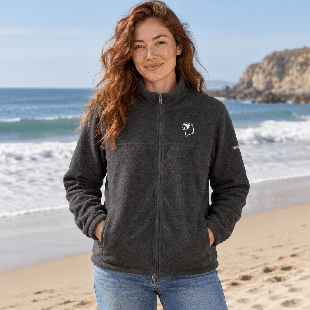 A woman with long wavy red hair stands on a sandy beach wearing a dark charcoal gray fleece zip-up jacket with a small white embroidered ParrotHead logo on the left chest, paired with blue jeans.