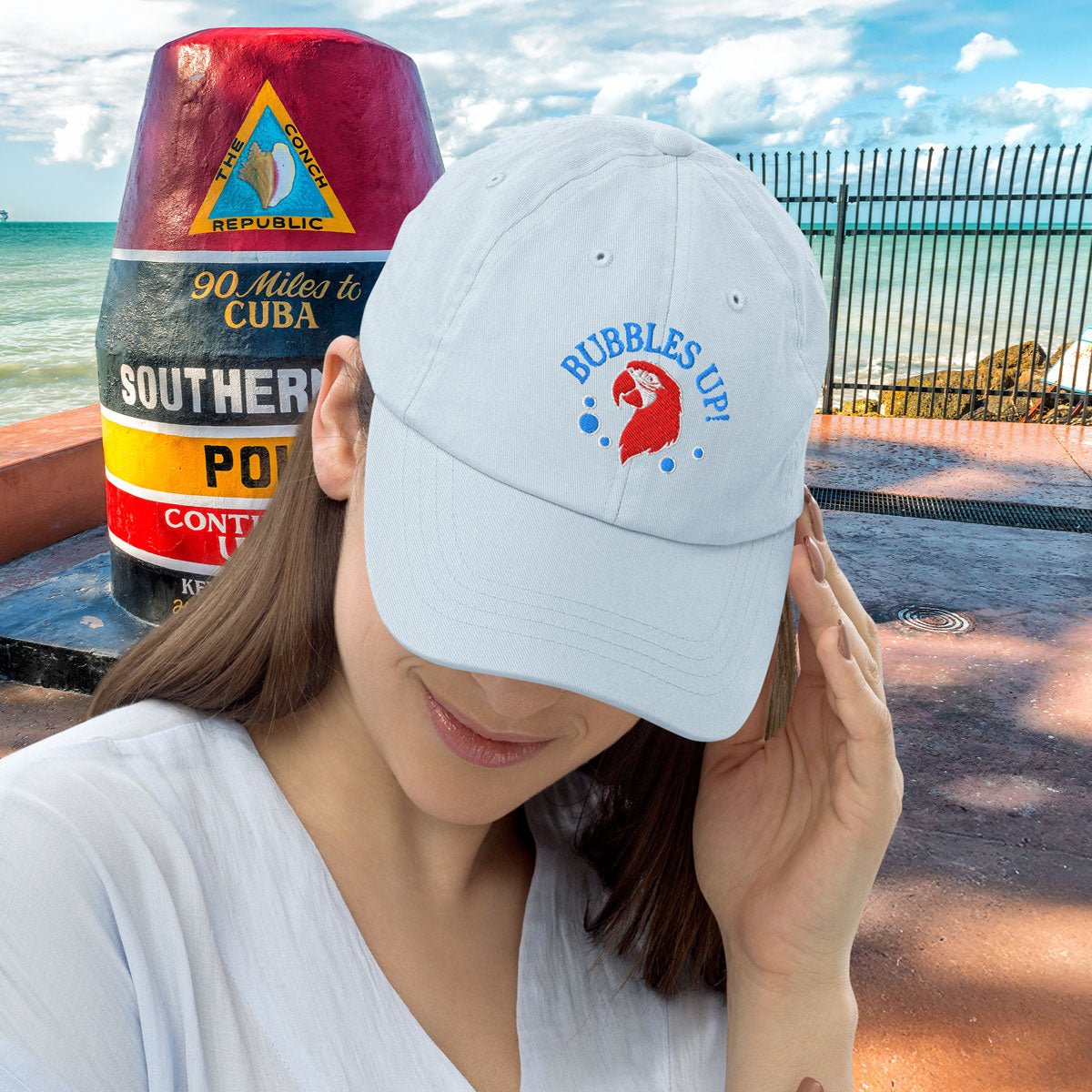 Woman wearing a pastel blue cap with 'Bubbles Up!' design, standing near a Southernmost point marker in Key West.