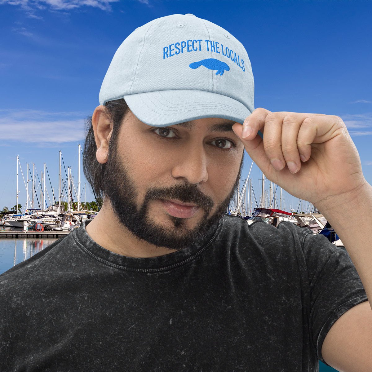 Man wearing a pastel blue Manatee Pastel Baseball Hat with 'Respect the Locals' text at a marina.