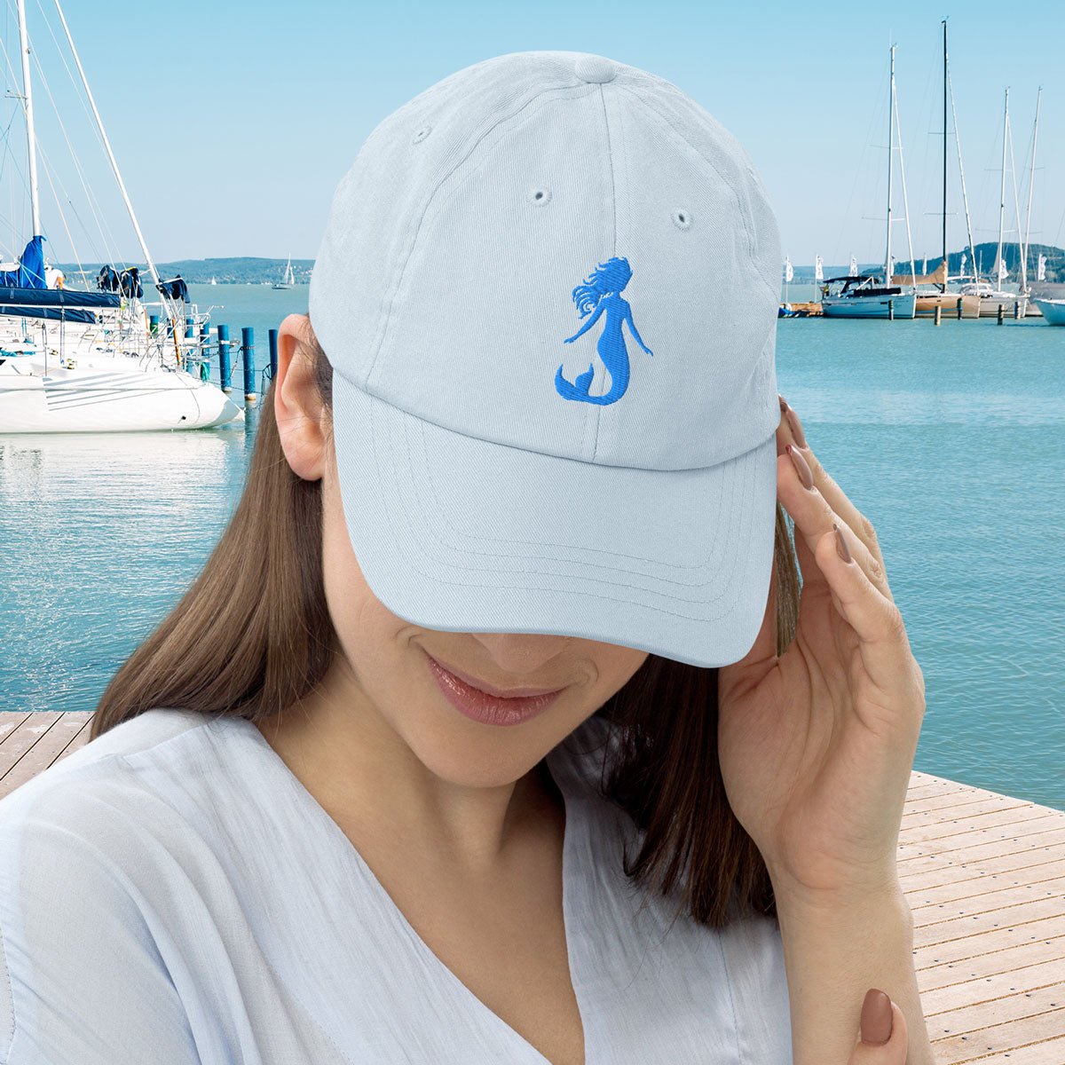 Woman wearing a light blue Pastel Mermaid baseball hat with a mermaid logo at a marina.