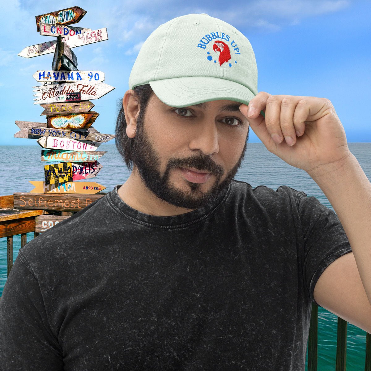 Man wearing a pastel mint cap with a bubbles up parrot head logo against a scenic background of Key West signpost.