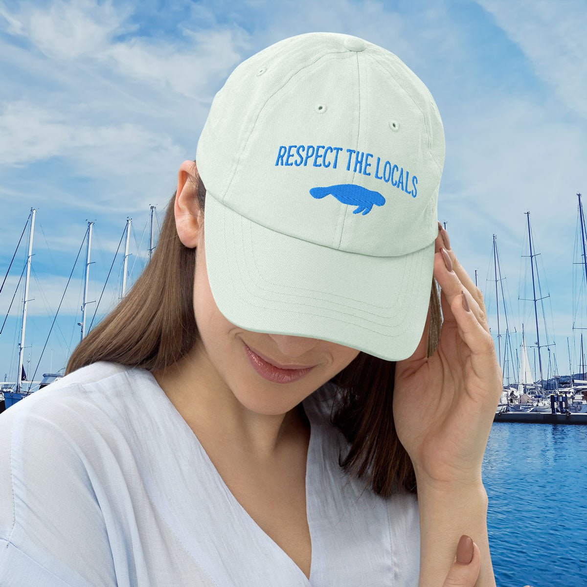 Woman wearing an embroidered manatee cap with 'Respect the Locals' text and a blue graphic, set against a marina background.