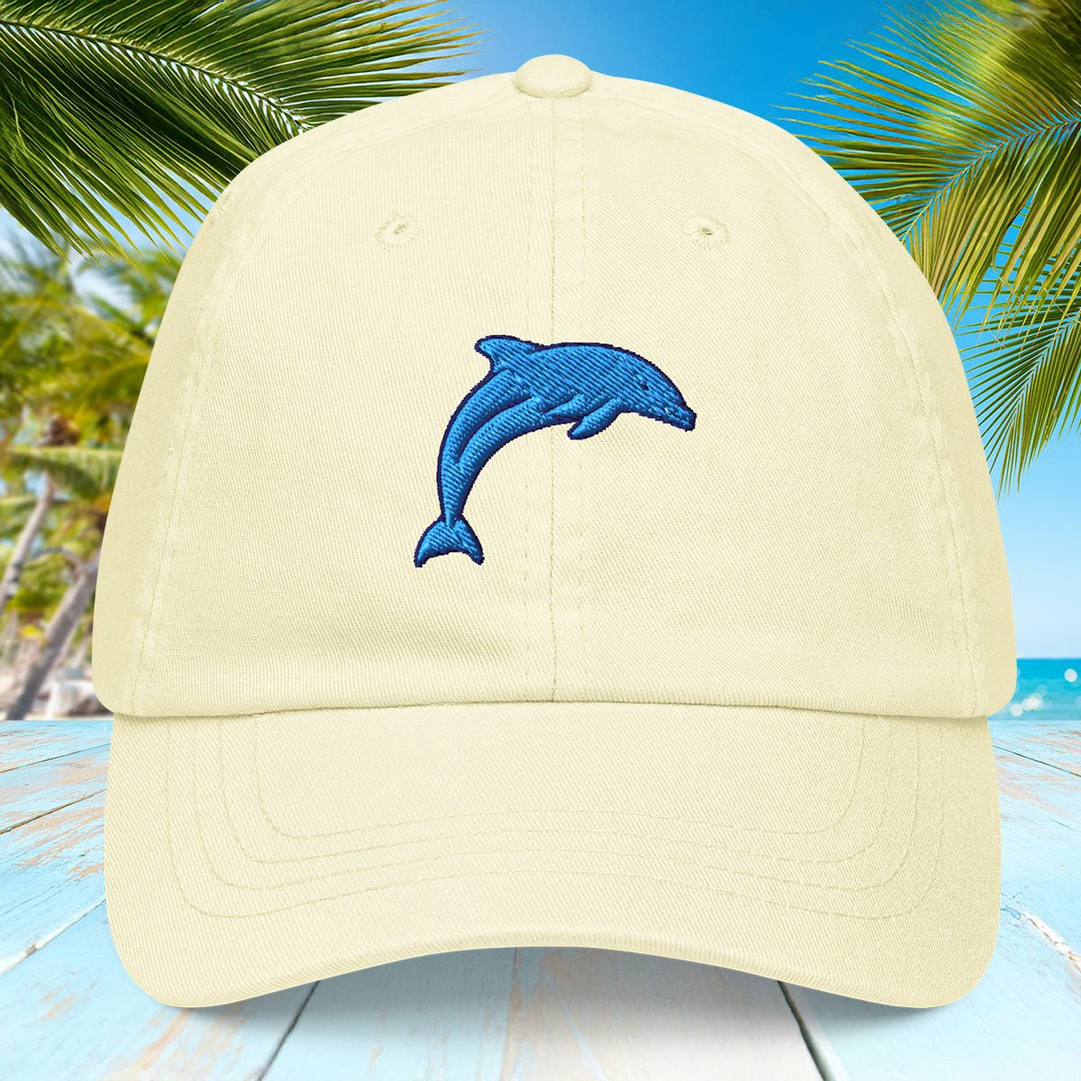Pastel yellow cap with a blue dolphin emblem on a tropical background with palm trees and blue water.