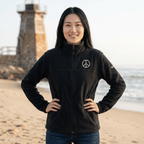 A woman stands on a sandy beach with the ocean and a lighthouse in the background, wearing a black zip-up fleece jacket with a white peace sign logo on the left chest.