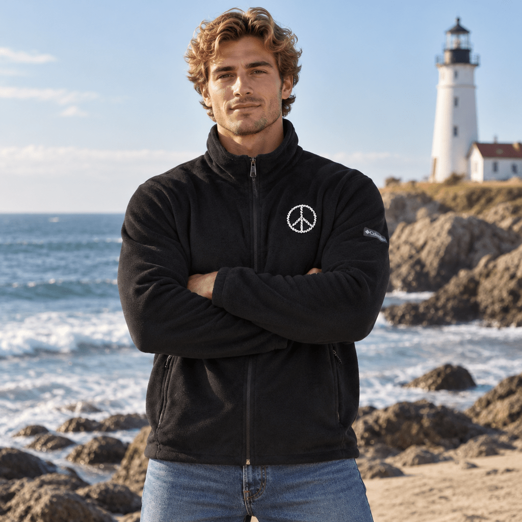 A man wearing a black fleece zip-up jacket with a white peace sign logo on the chest, standing on a rocky beach with the ocean and a lighthouse in the background.
