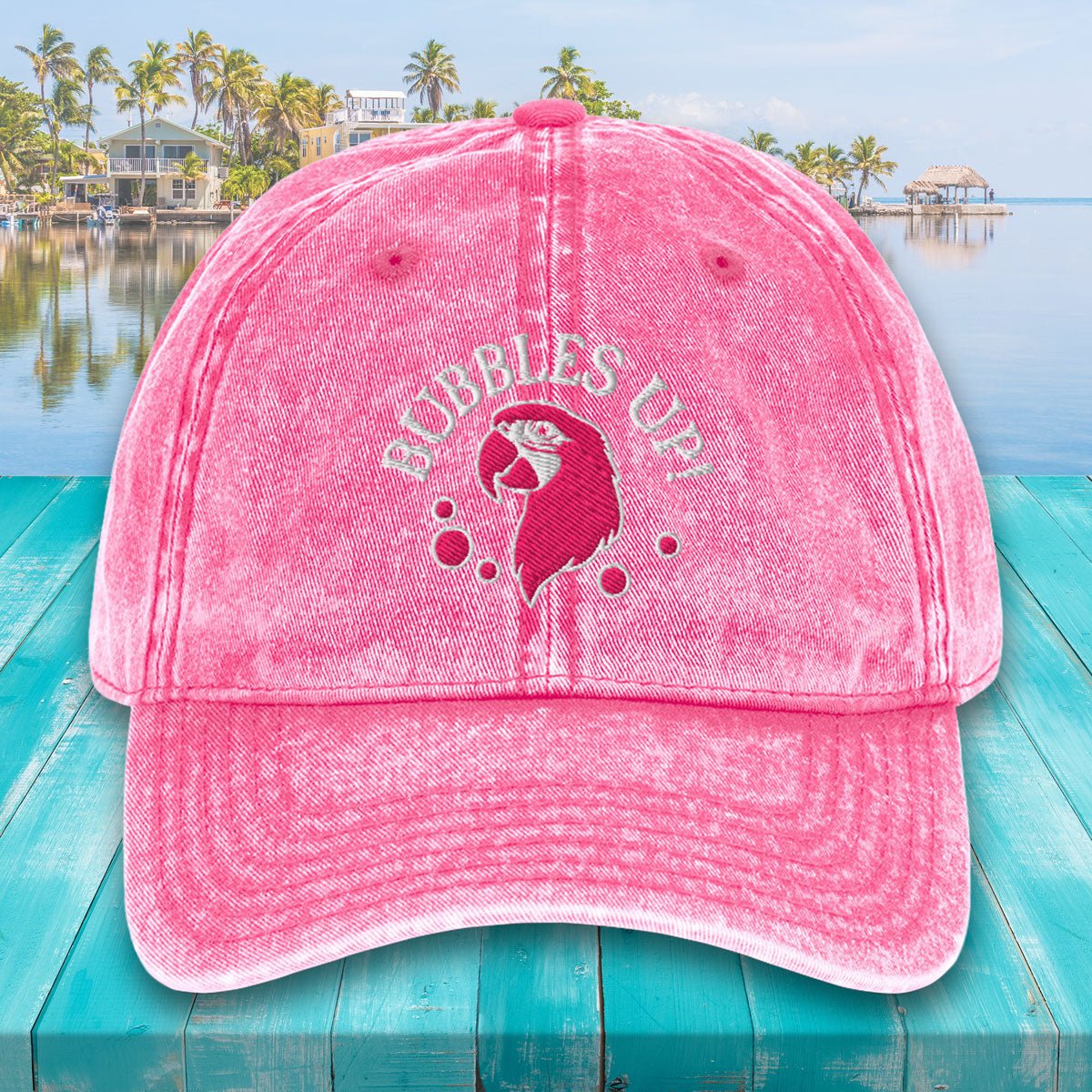 Pink cap with 'Bubbles Up!' text and mermaid design on a teal surface with a lake and palm trees in the background