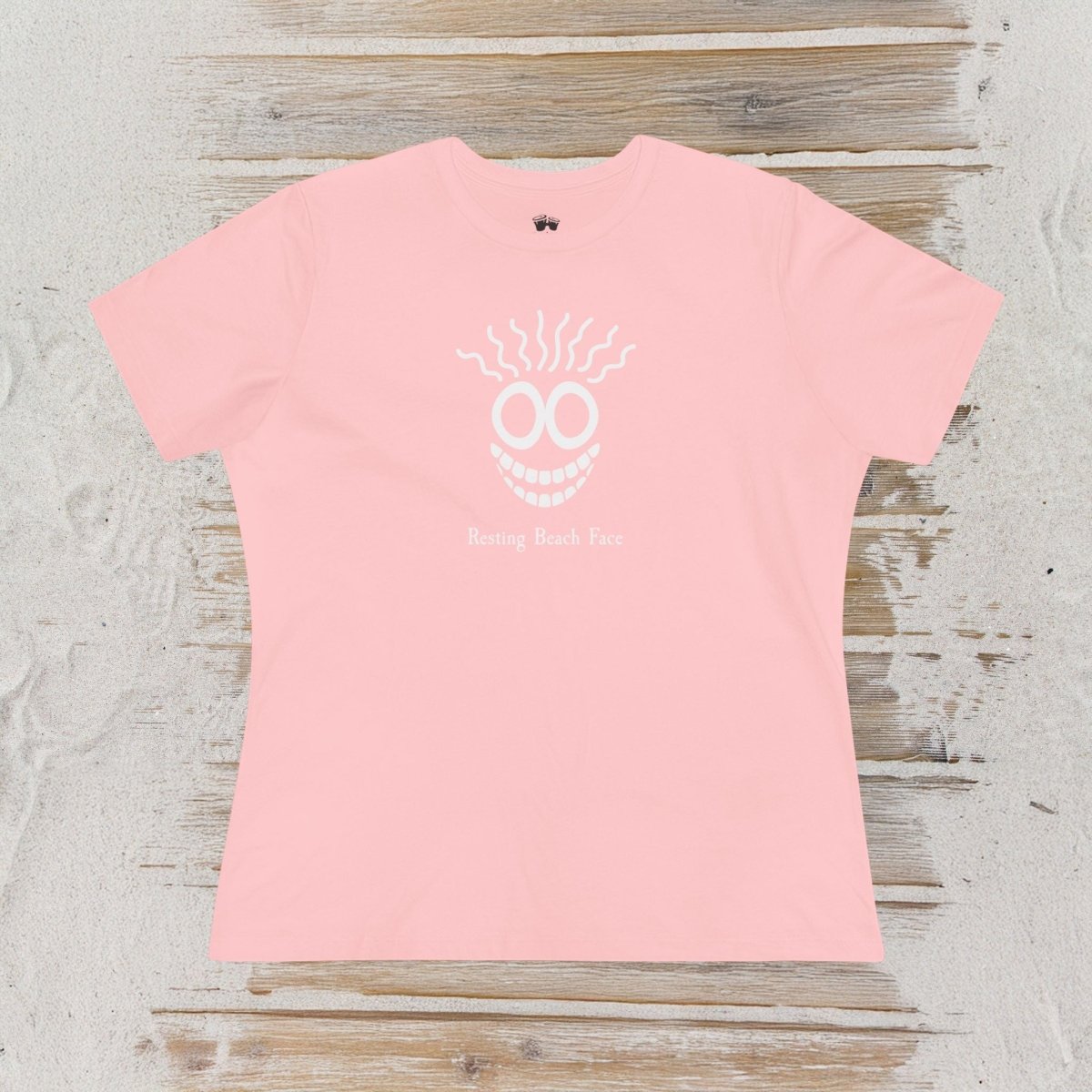 A light pink short-sleeve t-shirt with a white graphic of a smiling face with teeth and the text 'Being Book Face' underneath, laid flat on a rustic light wood background.