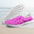 A pair of slip-on water shoes with a vibrant pink and magenta abstract pattern on the uppers and white soles, shown against a faded beach background.
