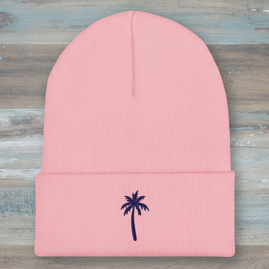 A light pink ribbed knit beanie hat with a folded cuff, featuring a small dark navy blue embroidered palm tree on the front, set against a plain white background.