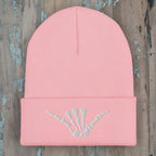 A light pink ribbed knit beanie hat with a folded cuff, featuring an embroidered white shaka sign skeleton hand design on the front.