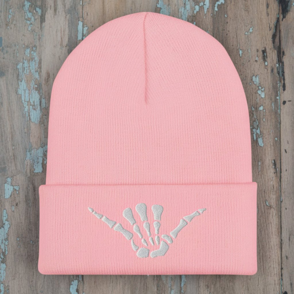 A light pink ribbed knit beanie hat with a folded cuff, featuring an embroidered white shaka sign skeleton hand design on the front.