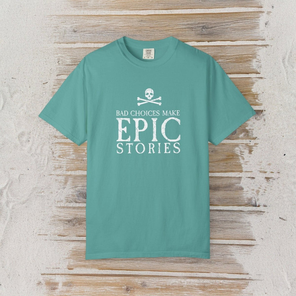 A teal short-sleeve crewneck t-shirt with a white graphic print on the front reading 'BAD CHOICES MAKE EPIC STORIES' below a skull and crossbones, displayed flat against a rustic light wood plank background.