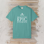 A teal short-sleeve crewneck t-shirt with a white graphic print on the front reading 'BAD CHOICES MAKE EPIC STORIES' below a skull and crossbones, displayed flat against a rustic light wood plank background.