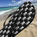 Black flip-flops with white skull and crossbones pattern on a white sandy beach background