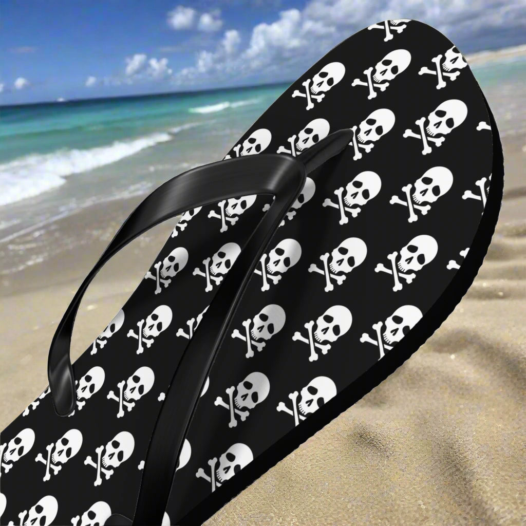 Black flip-flops with white skull and crossbones pattern on a white sandy beach background