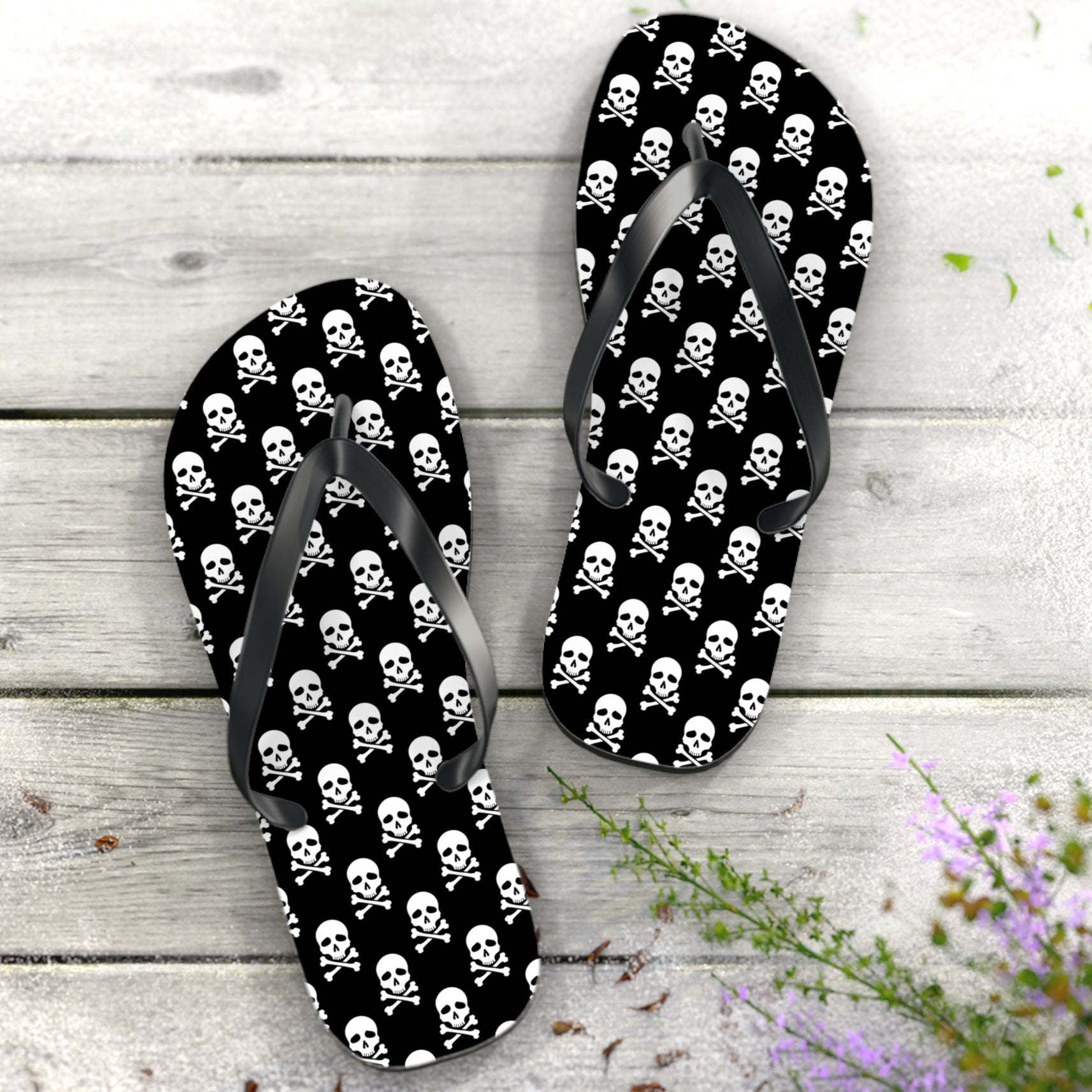 Flip-flops with skull and crossbones pattern on a wooden surface with flowers.