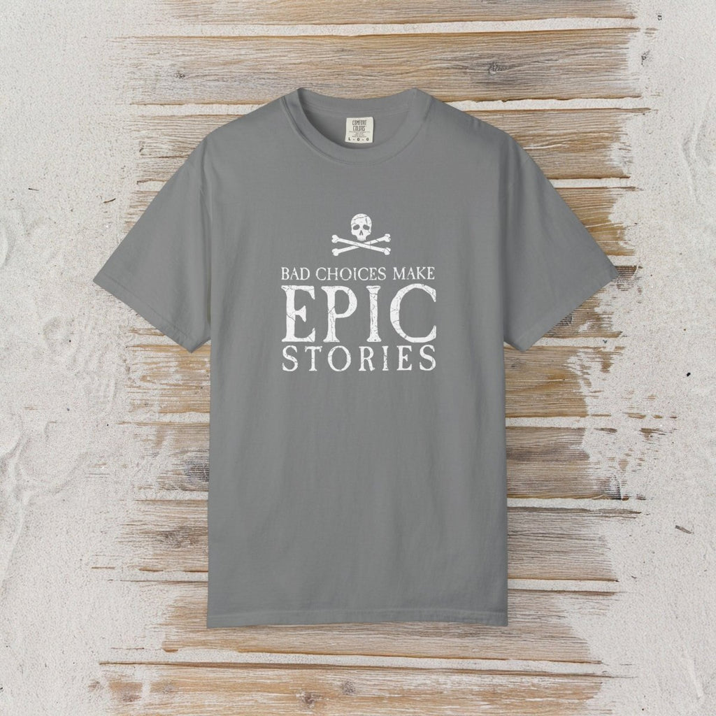 A gray short-sleeve Pirate T-Shirt with a white graphic print reading 'BAD CHOICES MAKE EPIC STORIES' and a skull and crossbones, laid flat on a rustic light wood background.