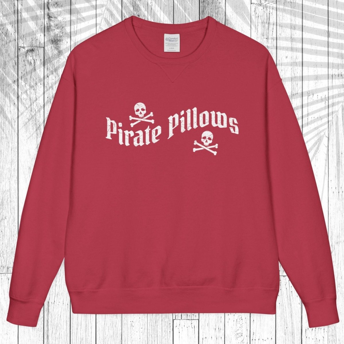 A red crewneck sweatshirt with 'Pirate Pillows' and skull-and-crossbones graphics on the chest, displayed flat against a white wood plank background.