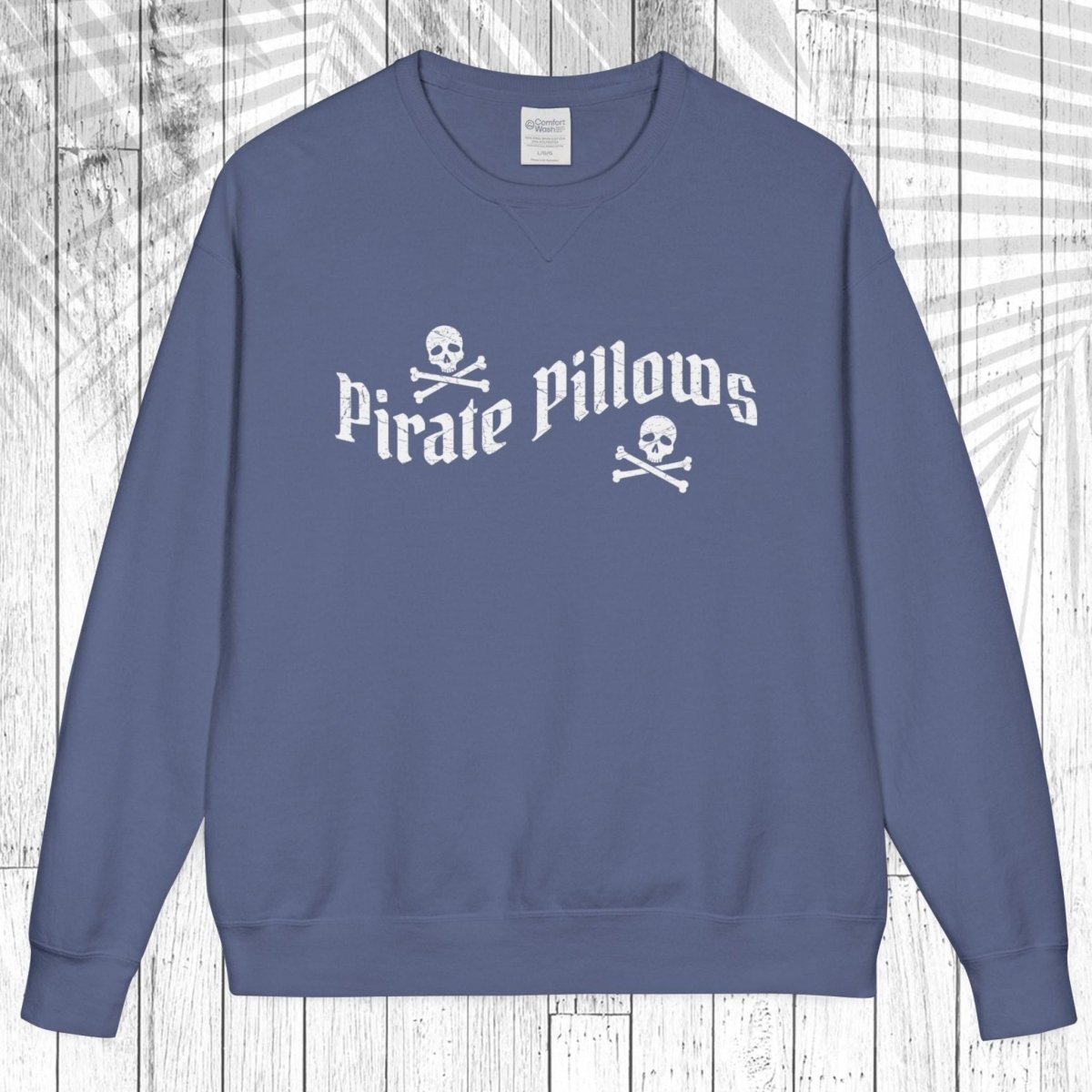 A slate blue crewneck sweatshirt featuring a white graphic print that reads 'Pirate Pillows' with skull and crossbones icons, displayed flat against a white wood plank background.