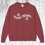 A crewneck sweatshirt in a dusty brick red color, featuring a white graphic print on the chest that reads 'Pirate Pillows' with skull and crossbones icons. The sweatshirt is displayed flat against a white wood plank background with a light gray palm leaf overlay.