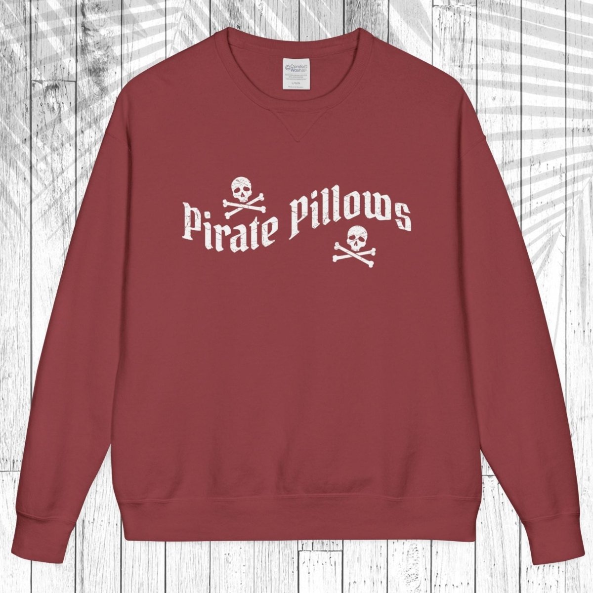 A crewneck sweatshirt in a dusty brick red color, featuring a white graphic print on the chest that reads 'Pirate Pillows' with skull and crossbones icons. The sweatshirt is displayed flat against a white wood plank background with a light gray palm leaf overlay.