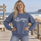 A woman smiling on a wooden boardwalk by the beach, wearing a slate blue crewneck sweatshirt with 'Pirate Pillows' and skull graphics printed in white across the chest.