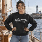 A woman wearing a black crewneck sweatshirt with a white 'Pirate Pillows' graphic stands on the deck of a sailboat with water and a lighthouse in the background.