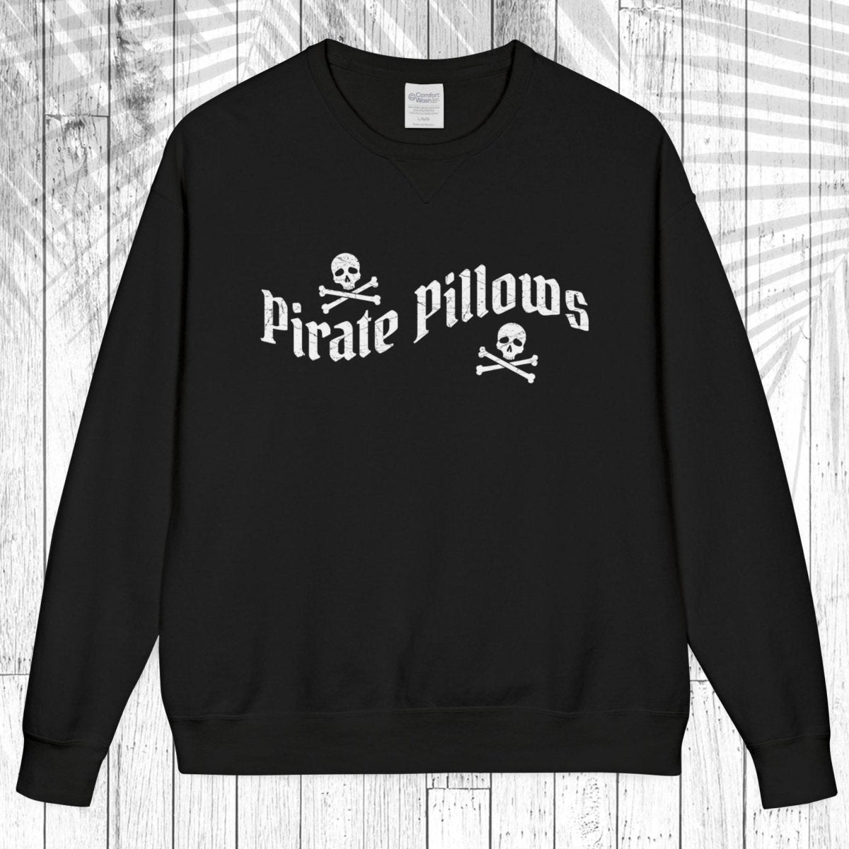 A black crewneck sweatshirt with a white graphic reading 'Pirate Pillows' and skull-and-crossbones icons, displayed flat against a white wood plank background.