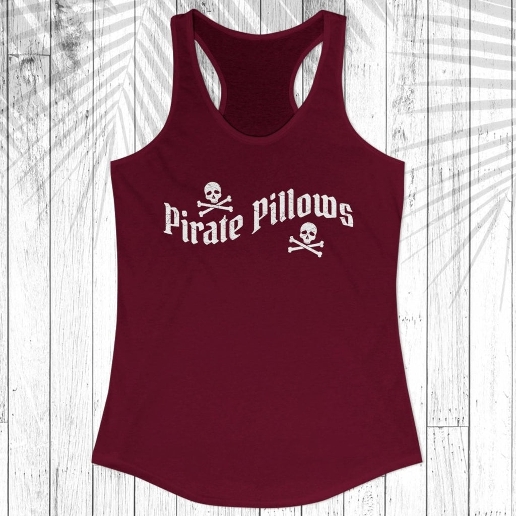 A women's racerback tank top in maroon with a white 'Pirate Pillows' text and skull-and-crossbones graphic on the chest, displayed flat against a white wood plank background with a palm leaf overlay.