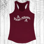 A women's racerback tank top in maroon with a white 'Pirate Pillows' text and skull-and-crossbones graphic on the chest, displayed flat against a white wood plank background with a palm leaf overlay.
