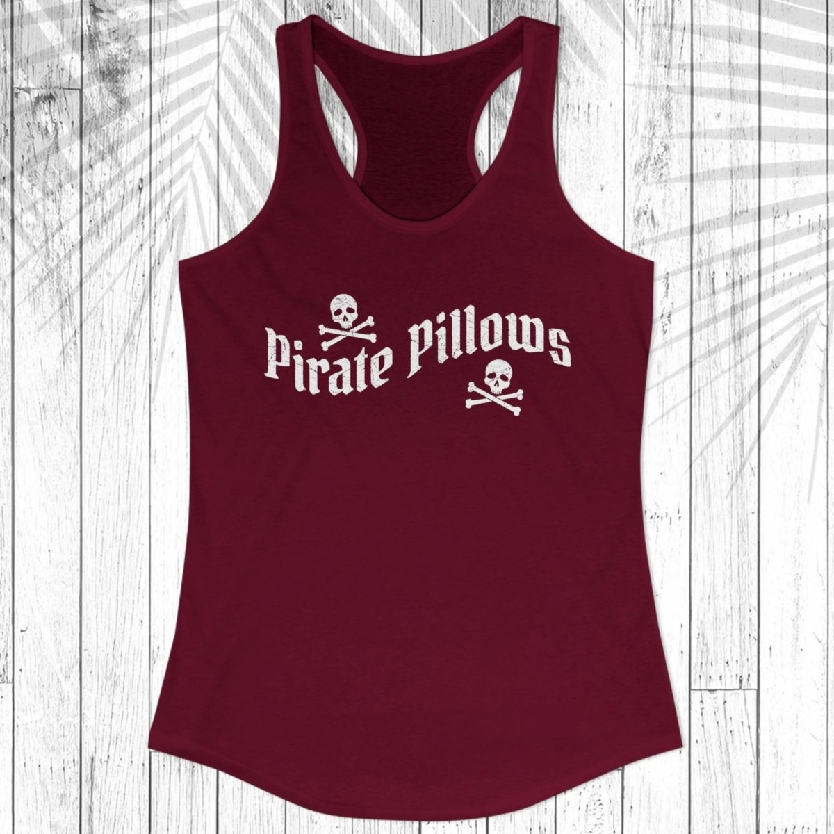 A women's racerback tank top in maroon with a white 'Pirate Pillows' text and skull-and-crossbones graphic on the chest, displayed flat against a white wood plank background with a palm leaf overlay.