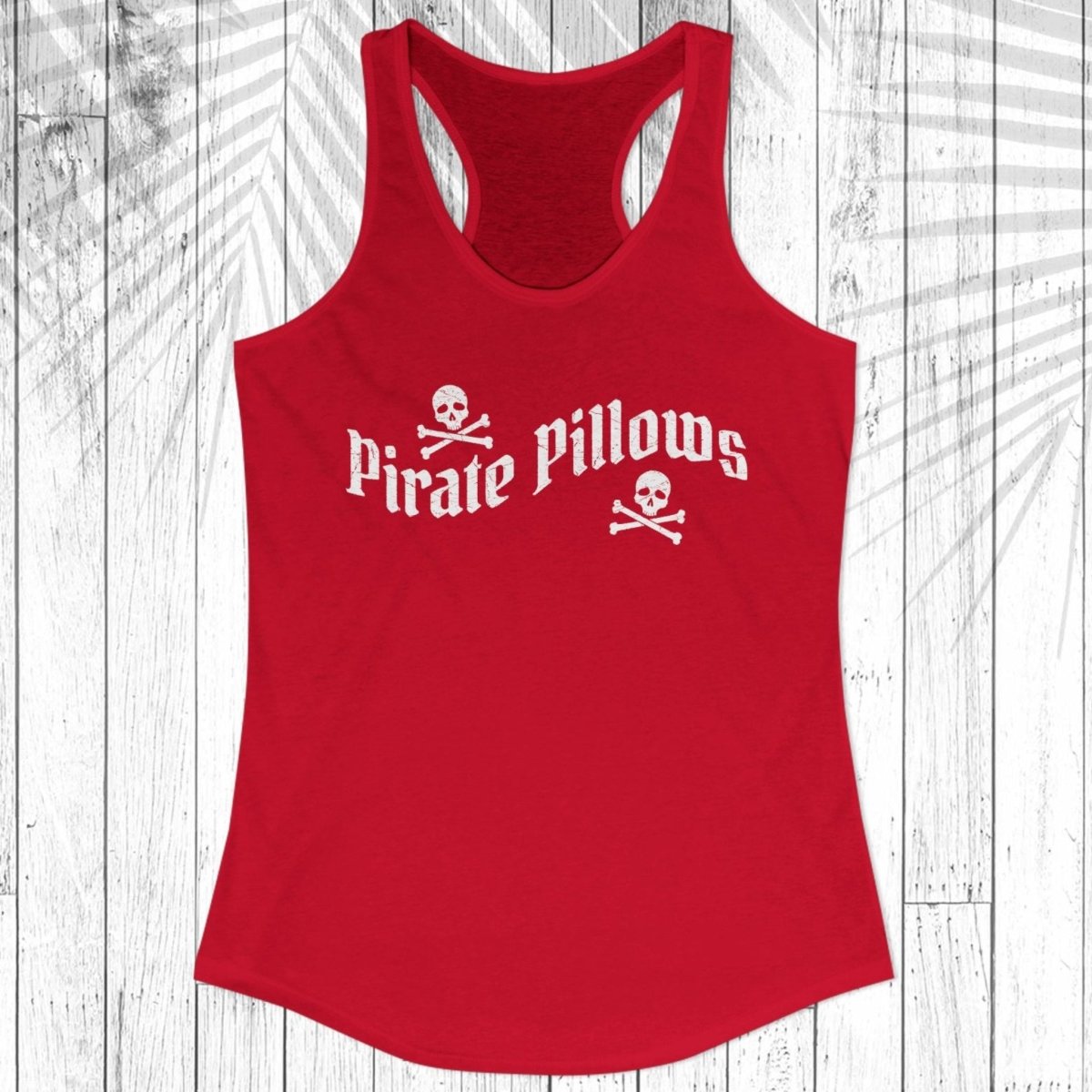 A red racerback tank top with 'Pirate Pillows' printed in white across the chest, displayed on a white wood plank background with a palm leaf overlay.