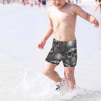 Child wearing skull-themed swim shorts playing in shallow water at the beach.