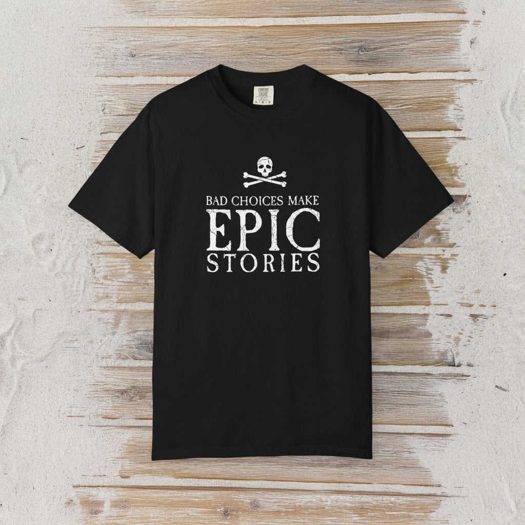 A black short-sleeve crewneck t-shirt with a white graphic print on the front reading 'BAD CHOICES MAKE EPIC STORIES' below a skull and crossbones, displayed flat against a rustic light wood plank background.
