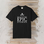 A black short-sleeve crewneck t-shirt with a white graphic print on the front reading 'BAD CHOICES MAKE EPIC STORIES' below a skull and crossbones, displayed flat against a rustic light wood plank background.