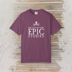 A short-sleeve crewneck t-shirt in a dusty plum purple color, featuring a white graphic print on the front with a skull and crossbones and the text 'BAD CHOICES MAKE EPIC STORIES'. The shirt is displayed flat against a rustic, whitewashed wooden plank background.